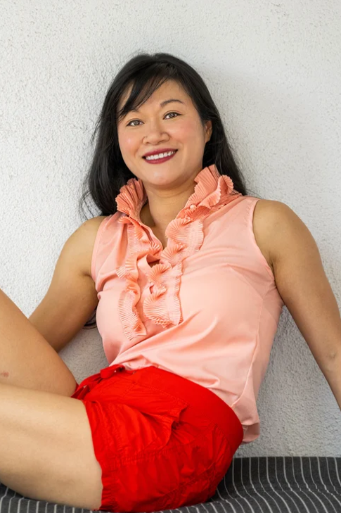 Portrait of a smiling woman in a peach blouse and red shorts, seated against a wall